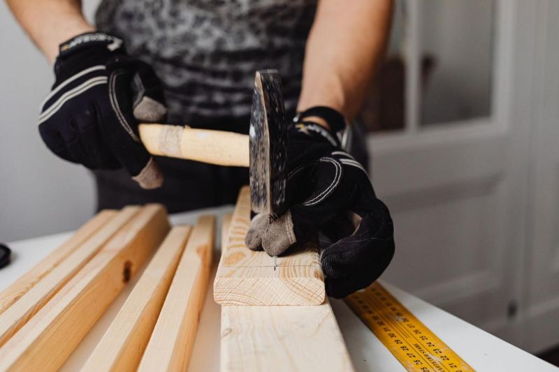 Manos con guantes clavando un clavo en un listón de madera